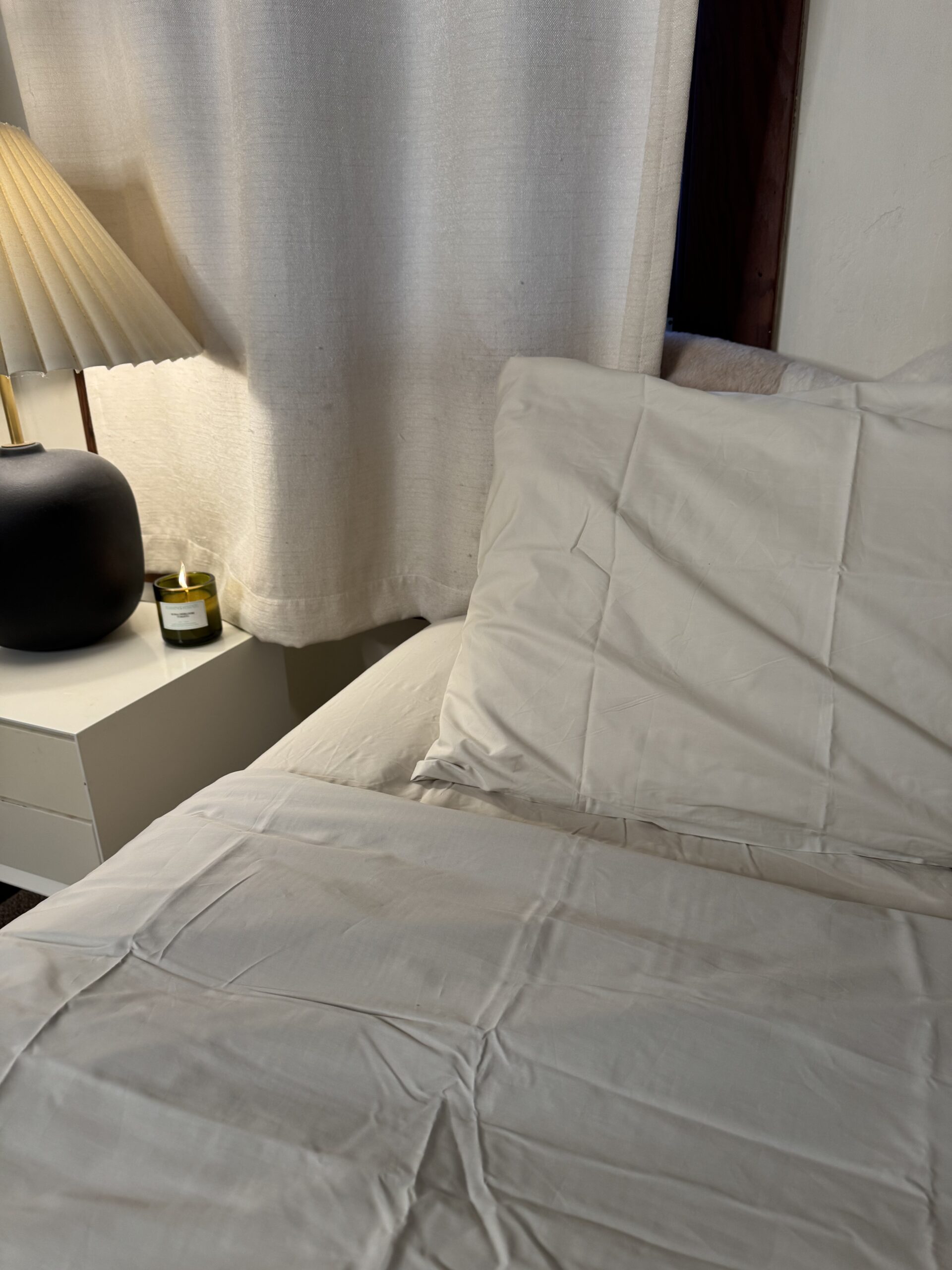 A neatly made bed with white sheets is next to a bedside table holding a lamp and a small candle. White curtains hang in the background.