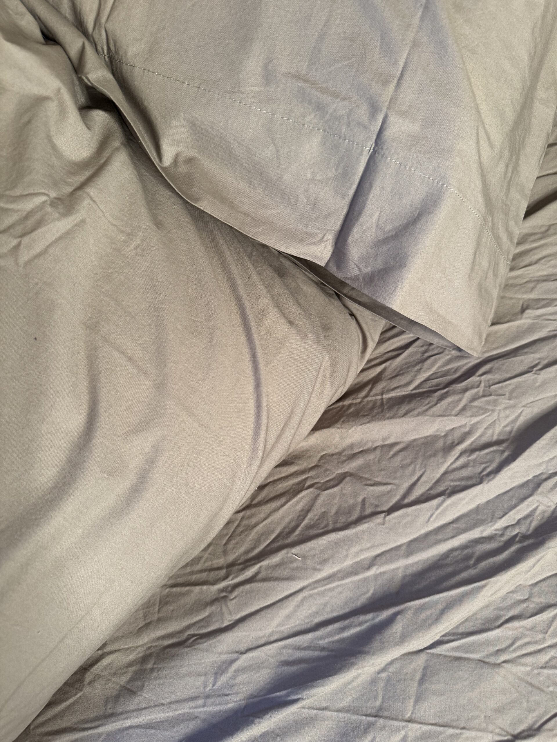 Wrinkled light gray bedding with a pillow on a mattress.