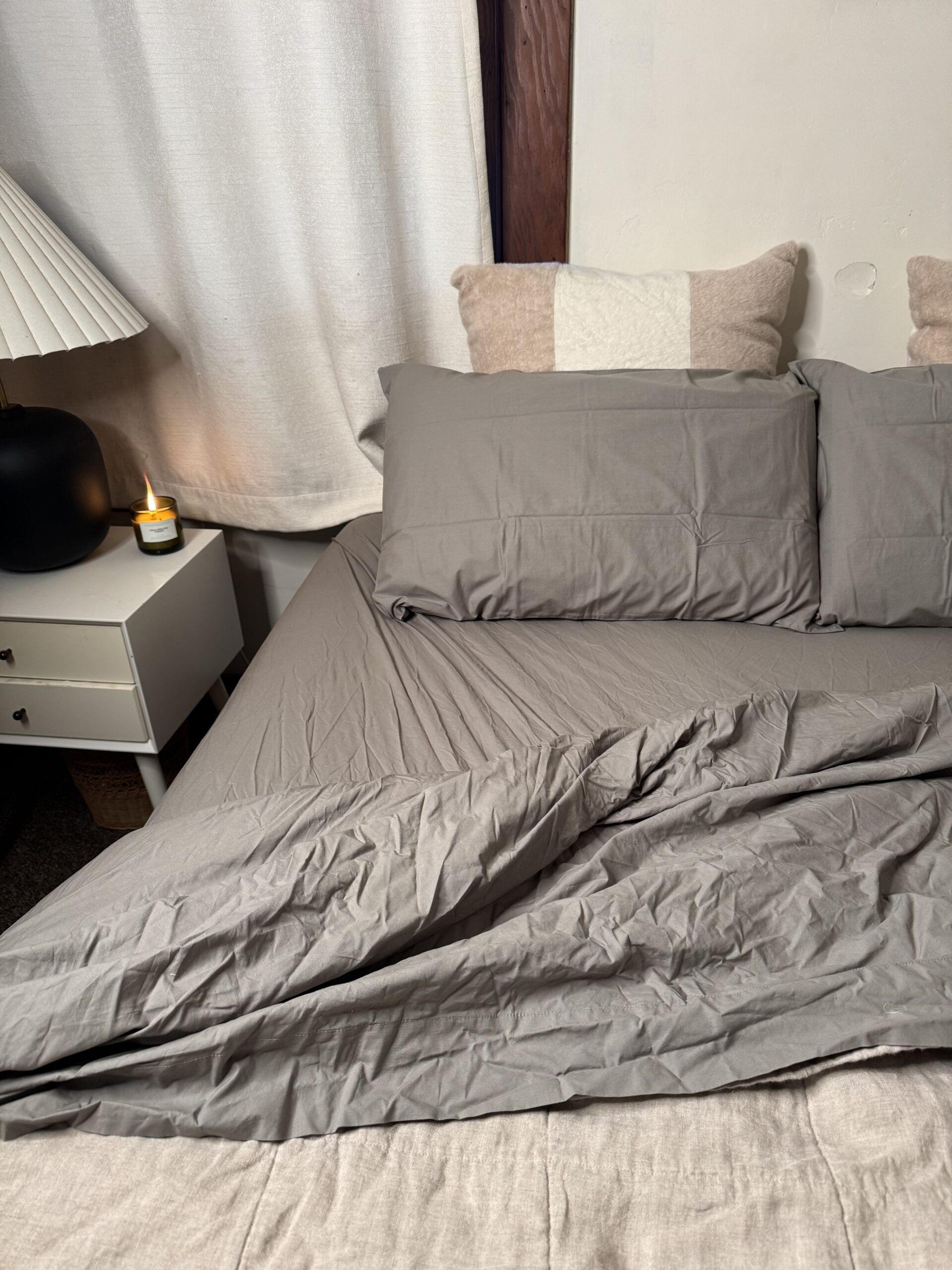 A neatly made bed with gray sheets and pillows, a white nightstand, a lamp, and a lit candle beside it.