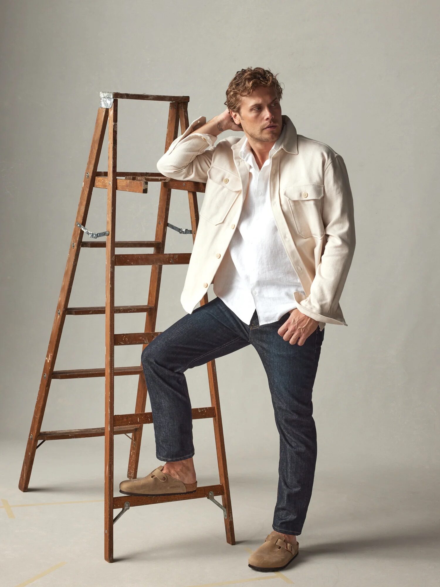 A person in a white jacket and jeans stands next to a wooden ladder, posing with one hand on their head.