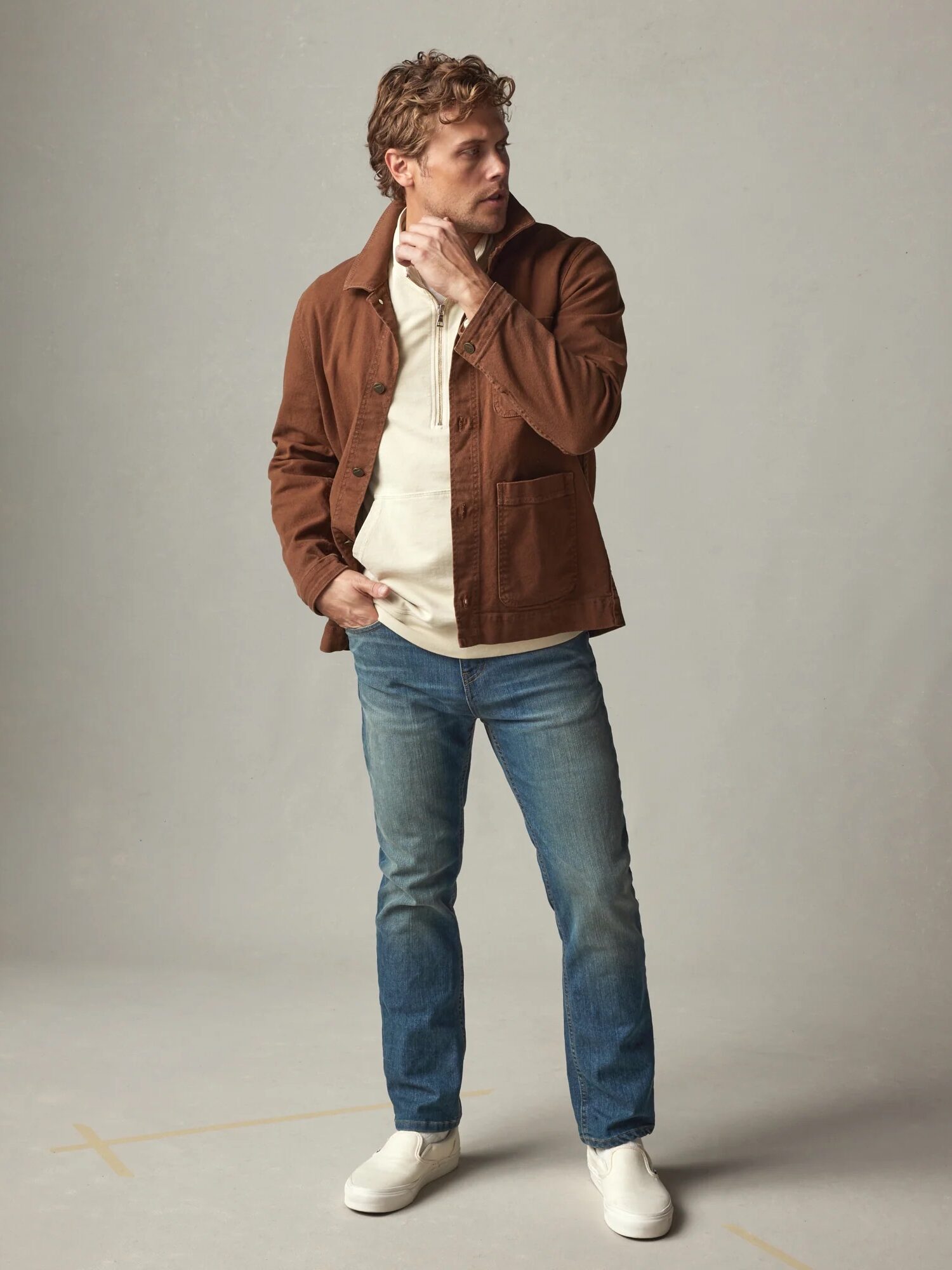 Man wearing a brown jacket, beige shirt, blue jeans, and white shoes, standing on a beige background.