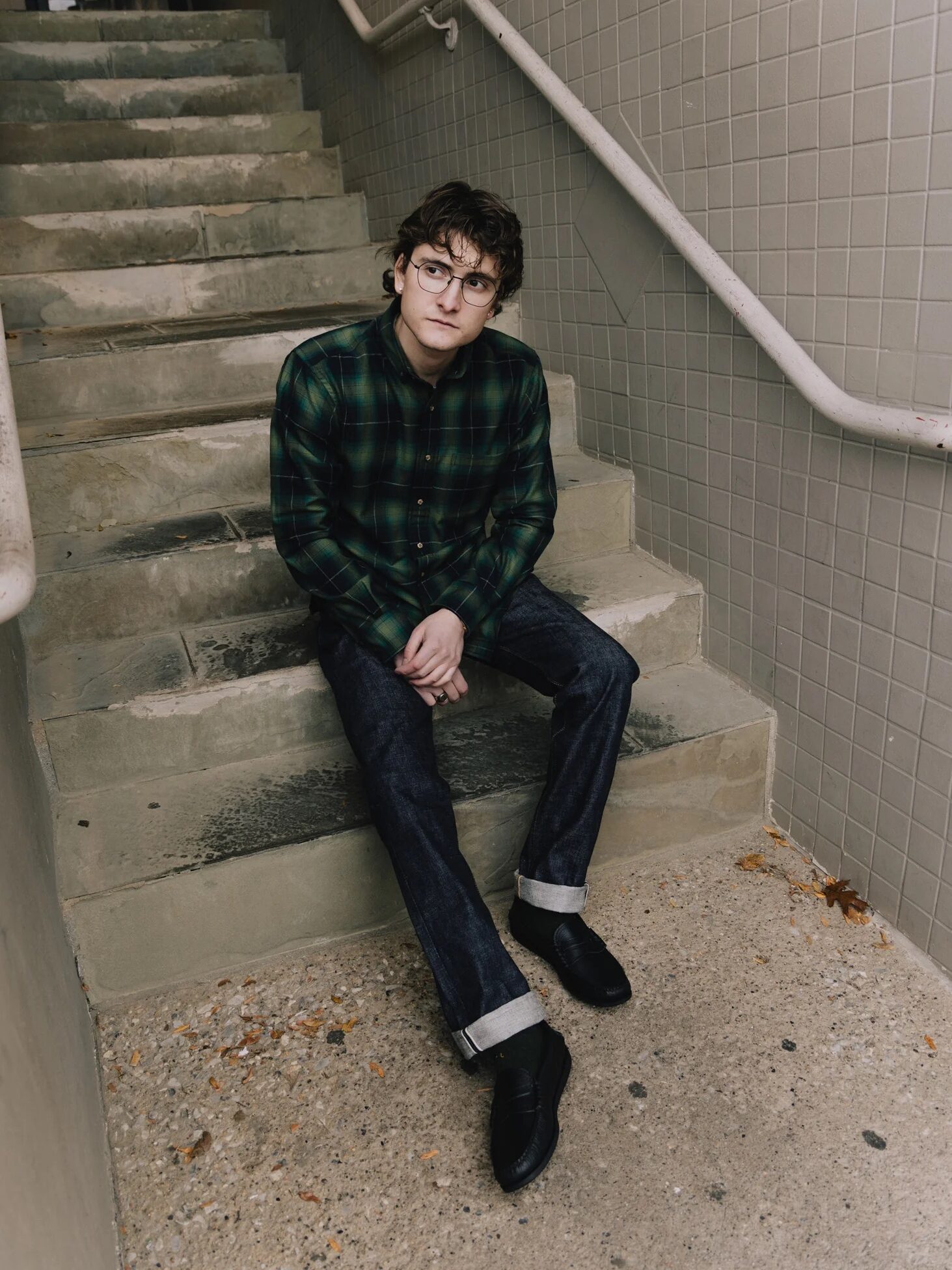 Person sitting on outdoor concrete steps, wearing glasses, a green plaid shirt, dark jeans, and black shoes.