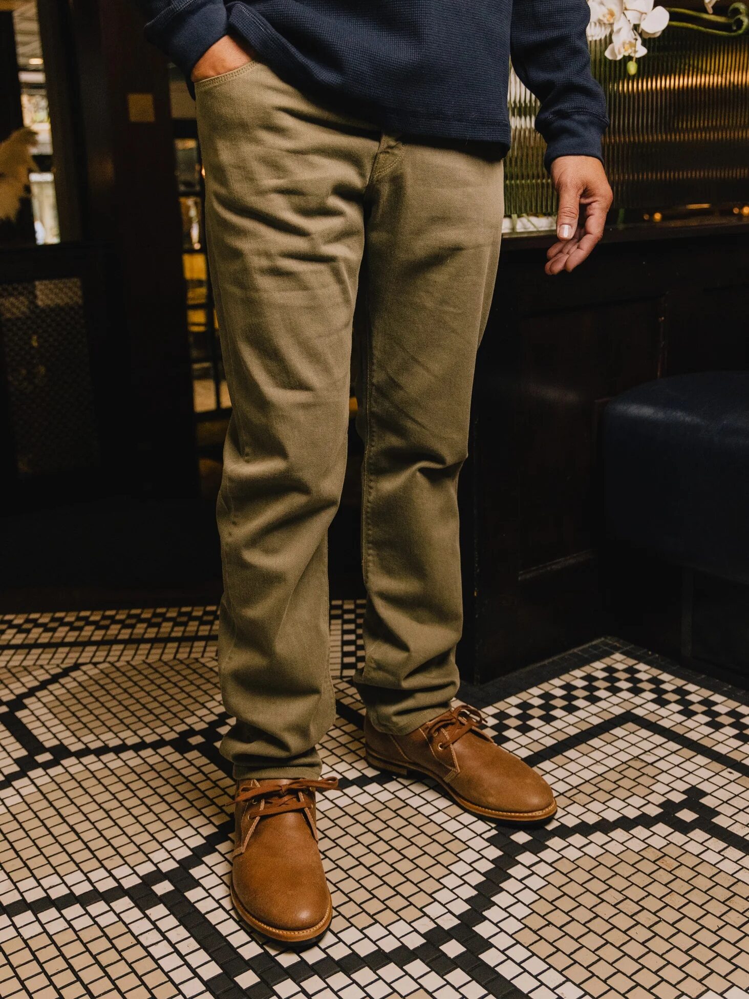 A person wearing olive green pants and brown leather shoes stands on a patterned tile floor.