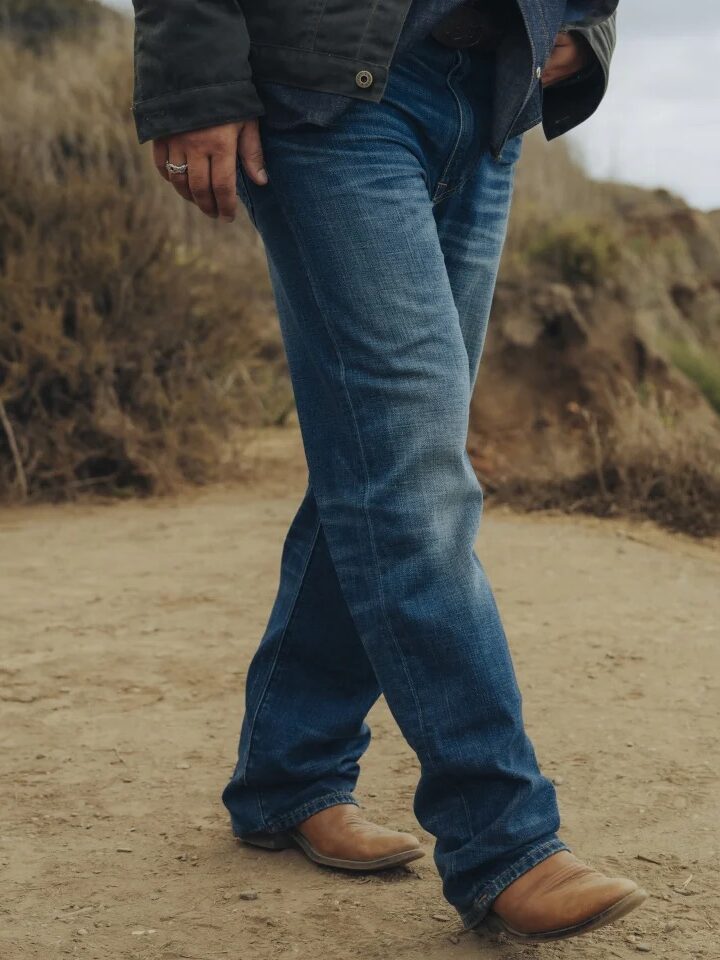 A person wearing blue jeans and brown cowboy boots walks on a dirt path near a rocky cliff.