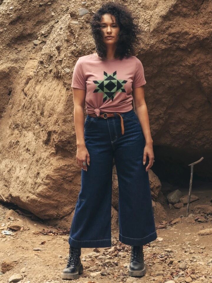 A person with curly hair is standing outdoors against a large rock, wearing a pink T-shirt with a geometric design and dark wide-legged jeans.