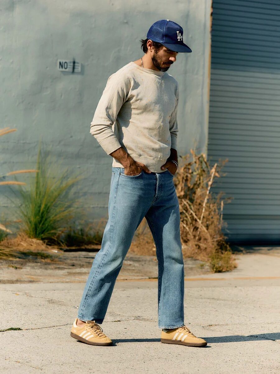 Man wearing a baseball cap, long-sleeve shirt, jeans, and sneakers walks on a sidewalk with hands in pockets. Background includes a corrugated metal door and dry plants.