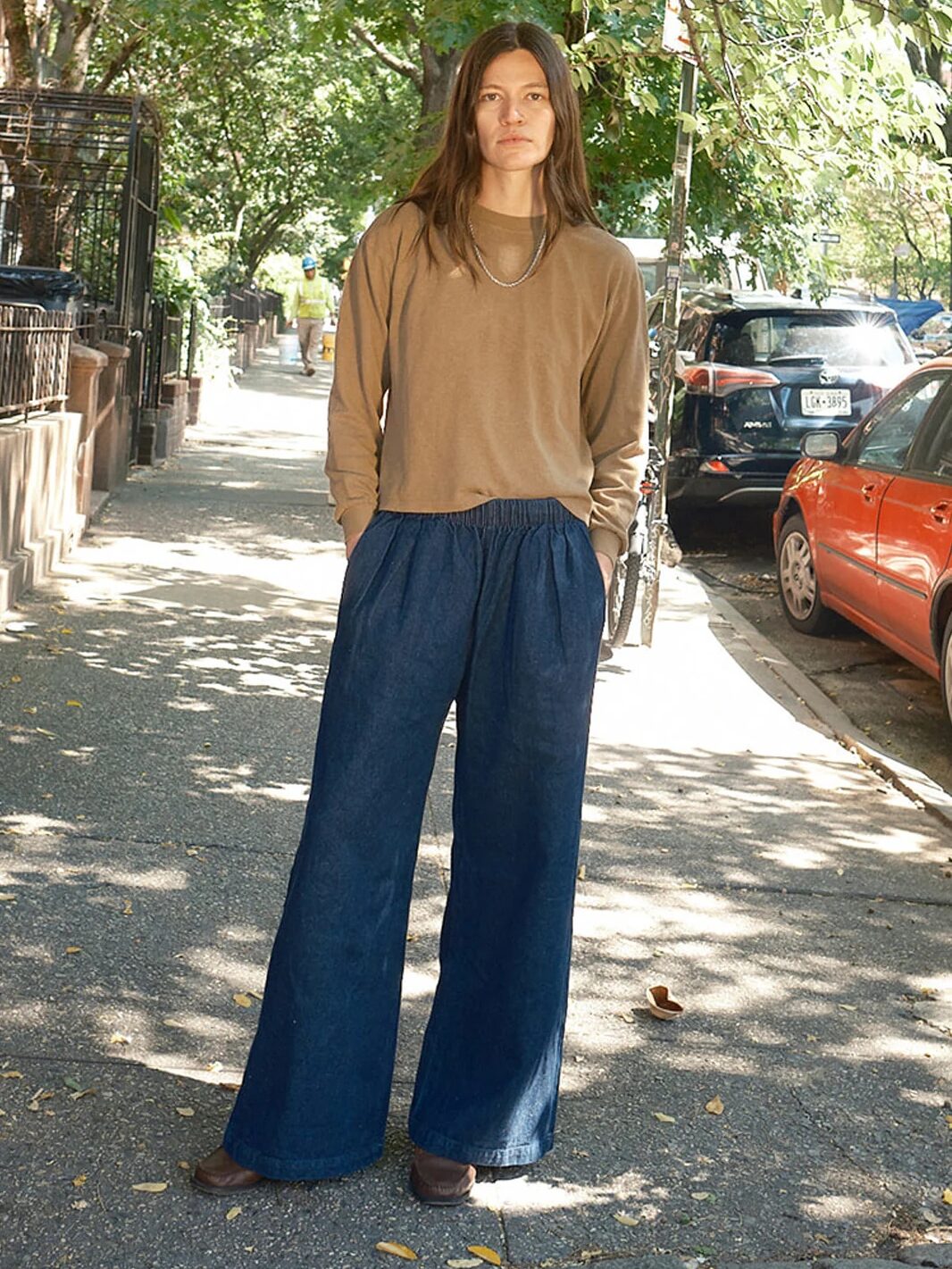 A person with long hair stands on a sidewalk wearing a brown long-sleeve shirt and wide-leg navy pants. Trees and parked cars line the street.