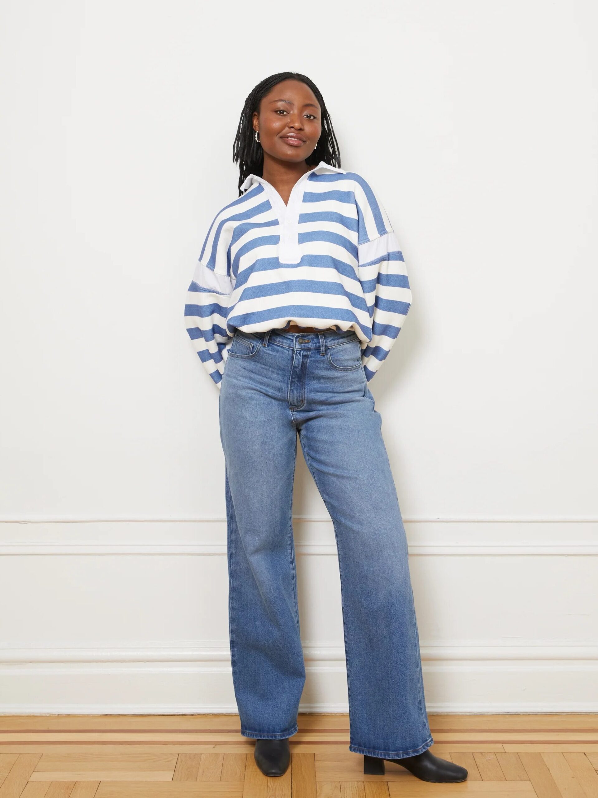 Person standing against a white wall, wearing a blue and white striped top, blue jeans, and black shoes.