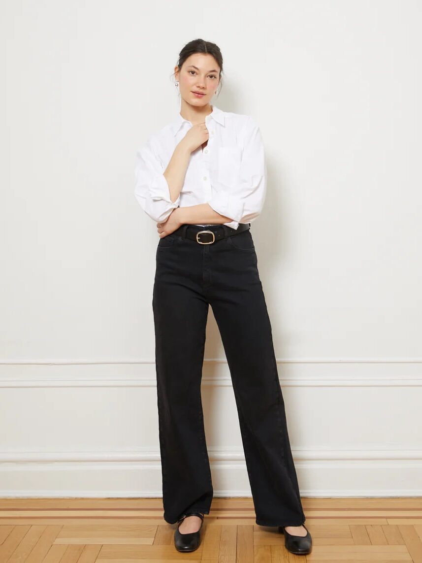 Person wearing a white shirt and black pants stands against a white wall, with one hand touching their face.