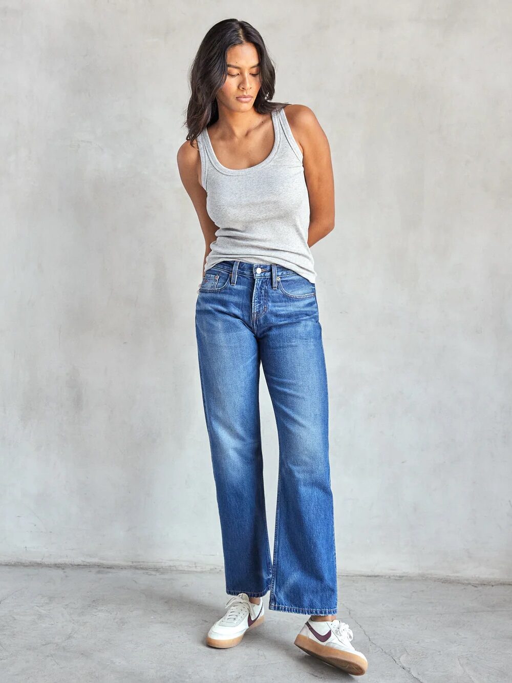 A person stands against a plain background wearing a gray tank top, blue jeans, and white sneakers, with hands behind their back.