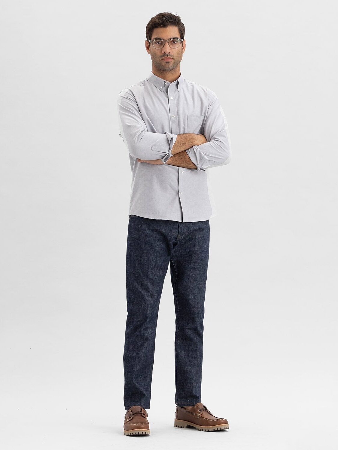 A man stands with arms crossed, wearing glasses, a light gray shirt, dark jeans, and brown shoes against a plain white background.