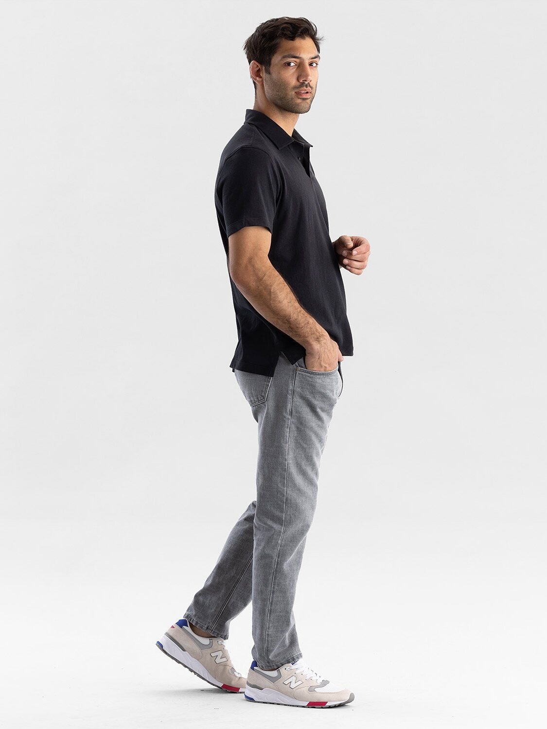 A man in a black polo and gray jeans stands against a plain white background, facing slightly to the side.