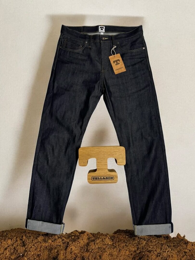 A pair of dark denim jeans with a folded cuff, displayed against a plain background. A cardboard tag is attached at the waist, and a wooden hanger with a "T" shape is beneath the jeans.