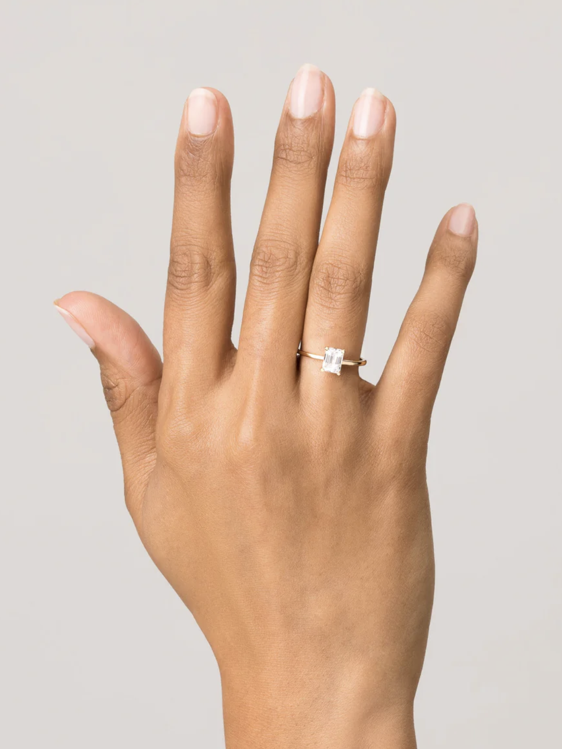 A hand wearing a gold ring with a square-cut gemstone on the ring finger, positioned against a plain background.