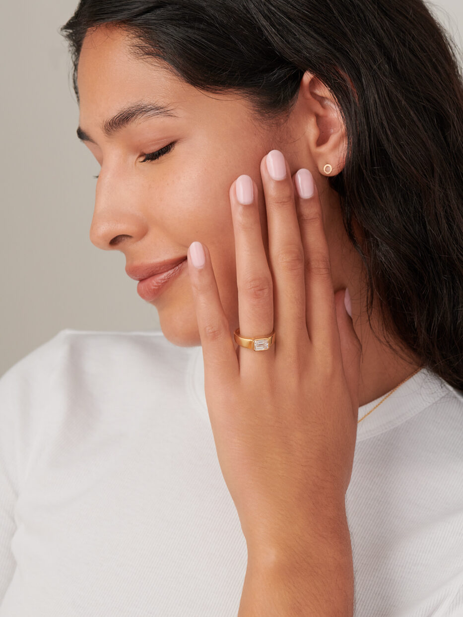 Person with long hair wearing a white top, displaying a gold ring and small earrings. One hand is touching the face. Eyes are closed.