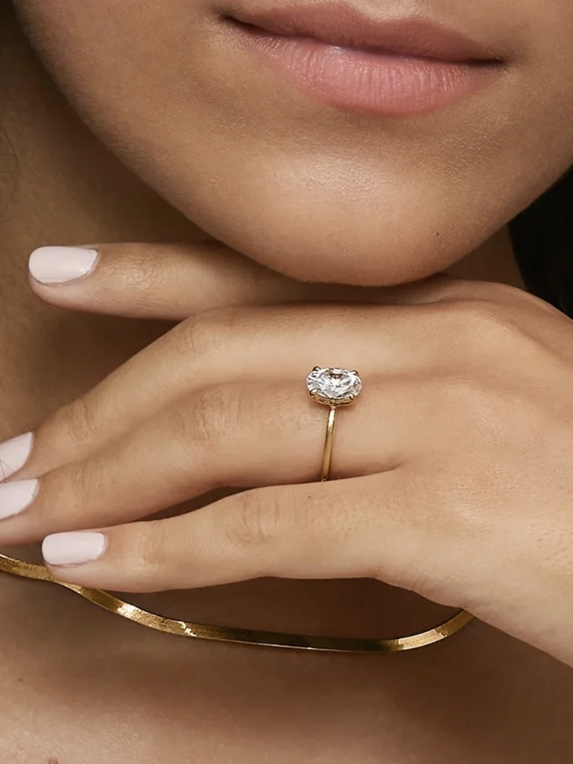 A person with neutral lipstick wears a gold oval ring on the ring finger and a thin gold necklace, with a minimalist style.