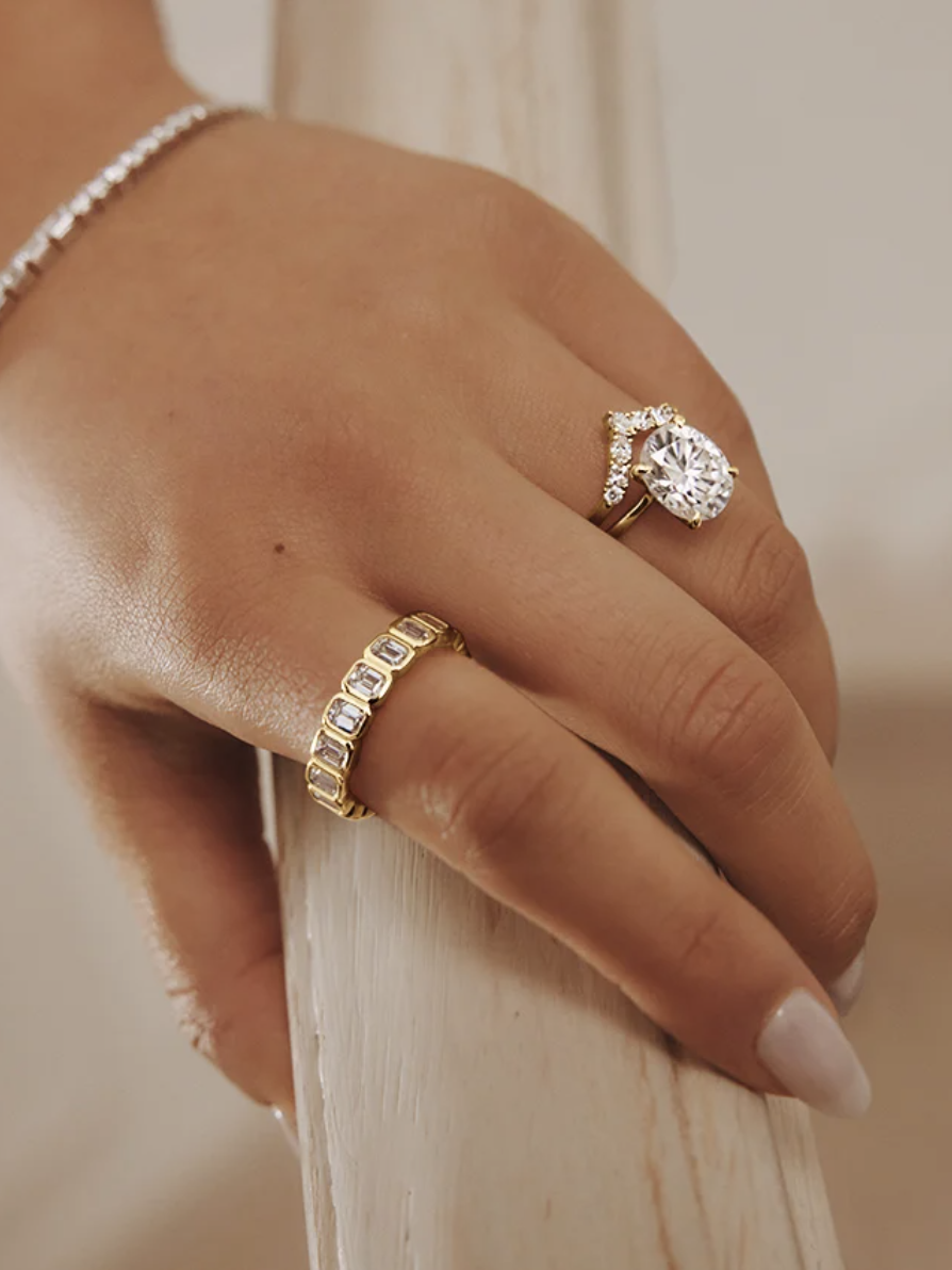 A hand wearing a diamond ring, a gold band with gems, and a silver bracelet rests on a railing.