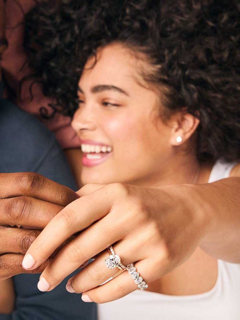 A smiling woman with curly hair displays her hand adorned with a diamond engagement ring and wedding band.