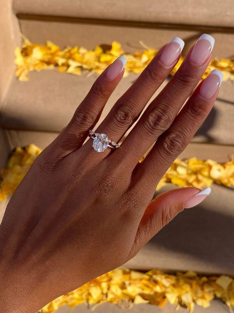 A hand with a French manicure displays a large diamond ring, surrounded by scattered yellow petals on a staircase.