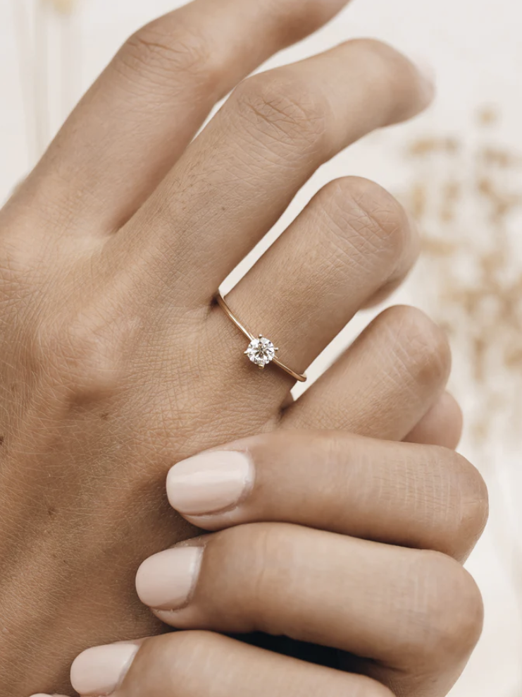 A hand wearing a simple gold ring with a single diamond is shown against a soft, neutral background.