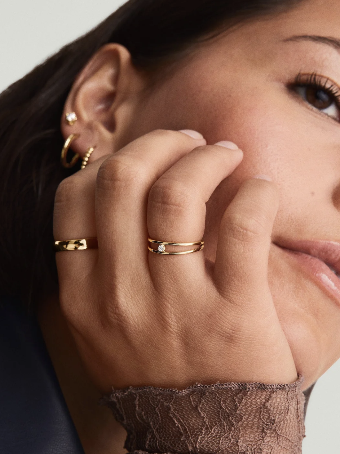 Close-up of a person touching their face, showcasing multiple gold rings on their fingers and gold hoop earrings. The lace sleeve adds a textured detail.