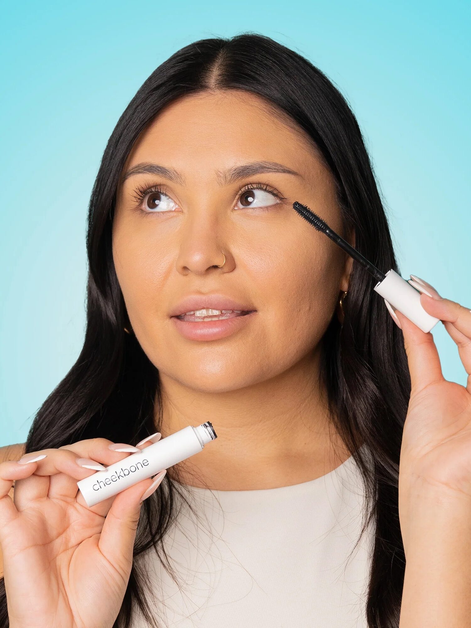 Person holding a mascara wand and tube, looking upwards. Light blue background.