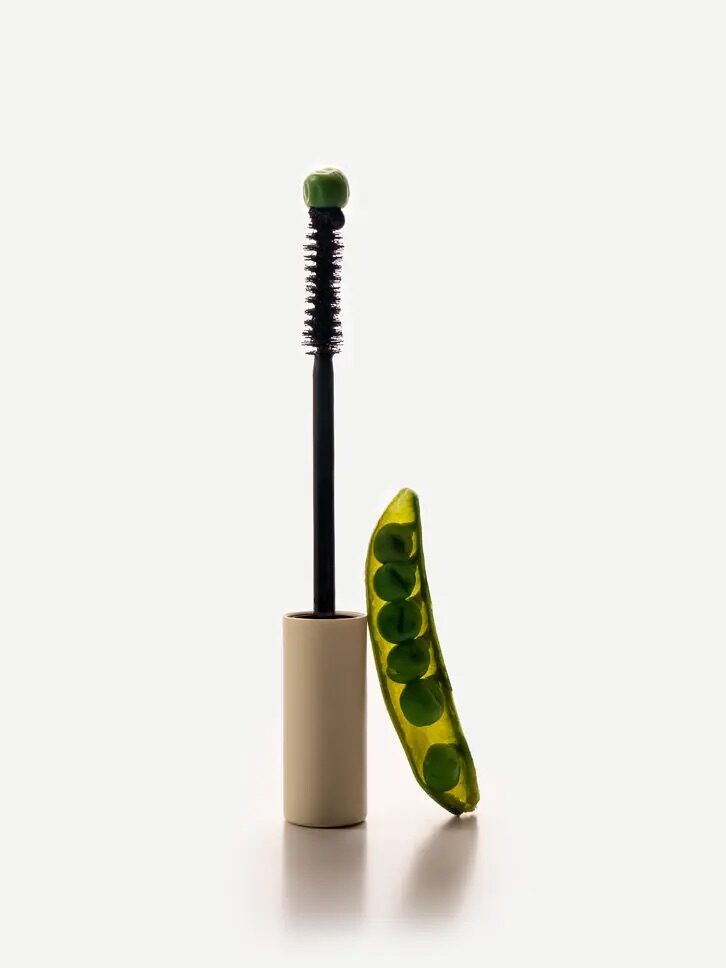 A mascara wand with a pea on top, standing next to an open pea pod containing several peas, against a plain background.