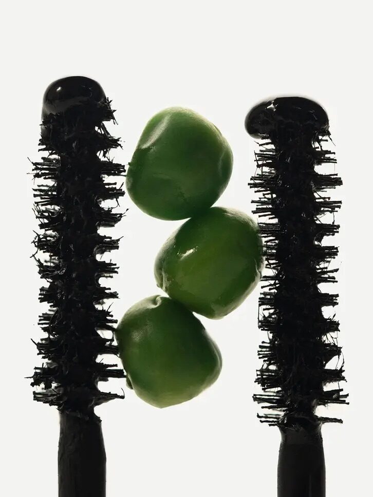 Two mascara brushes with green peas stacked between them on a white background.