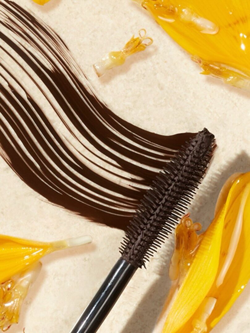 A mascara wand with dark brown mascara is laid on a light surface, surrounded by yellow flower petals and stamens.