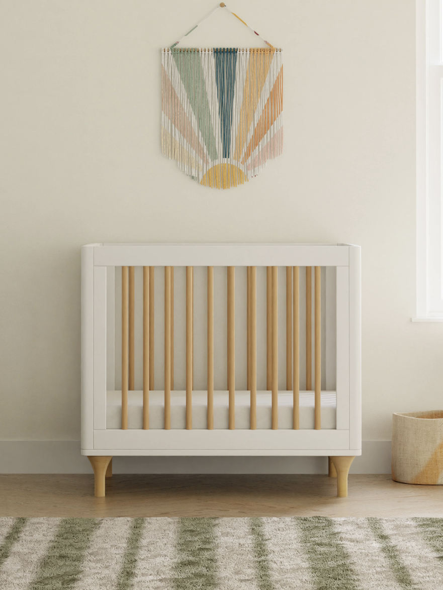 A minimalist nursery with a white and wood crib, a dresser to the left, a geometric wall hanging, a woven basket, and a striped rug on the floor.