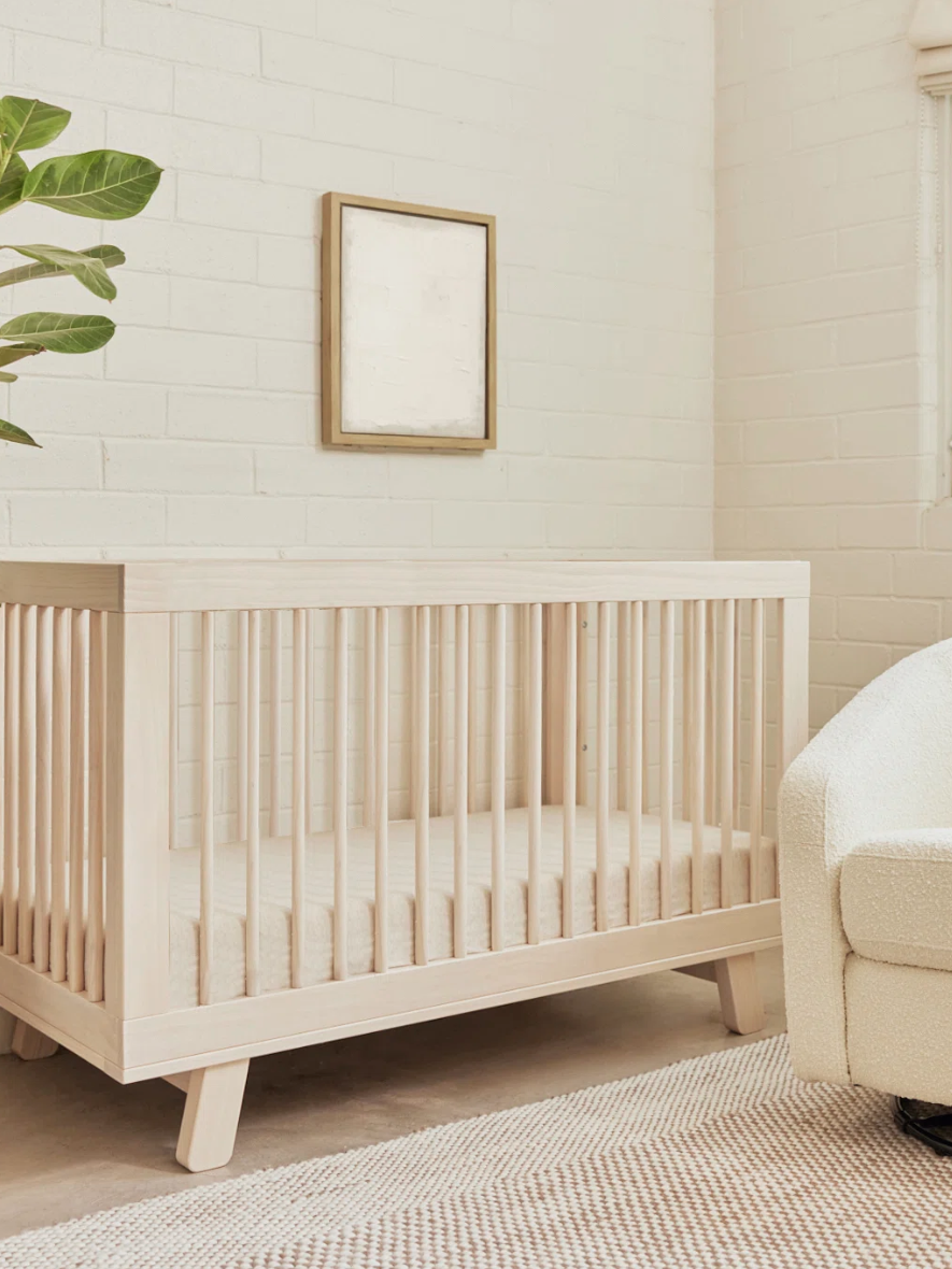 Minimalist nursery with a wooden crib, an armchair, a potted plant, and a window. Walls are white brick with a framed picture. Natural light fills the room.