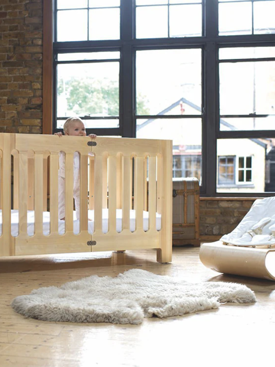 A baby stands in a wooden crib in a spacious room with large windows. A sheepskin rug and baby chair are on the wooden floor.