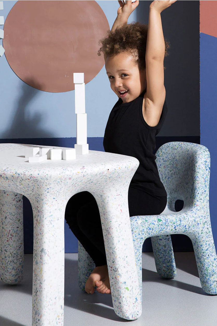 Child sitting on a speckled chair at a matching table, excitedly raising arms, with a small white block tower in front. Geometric wall art in background.