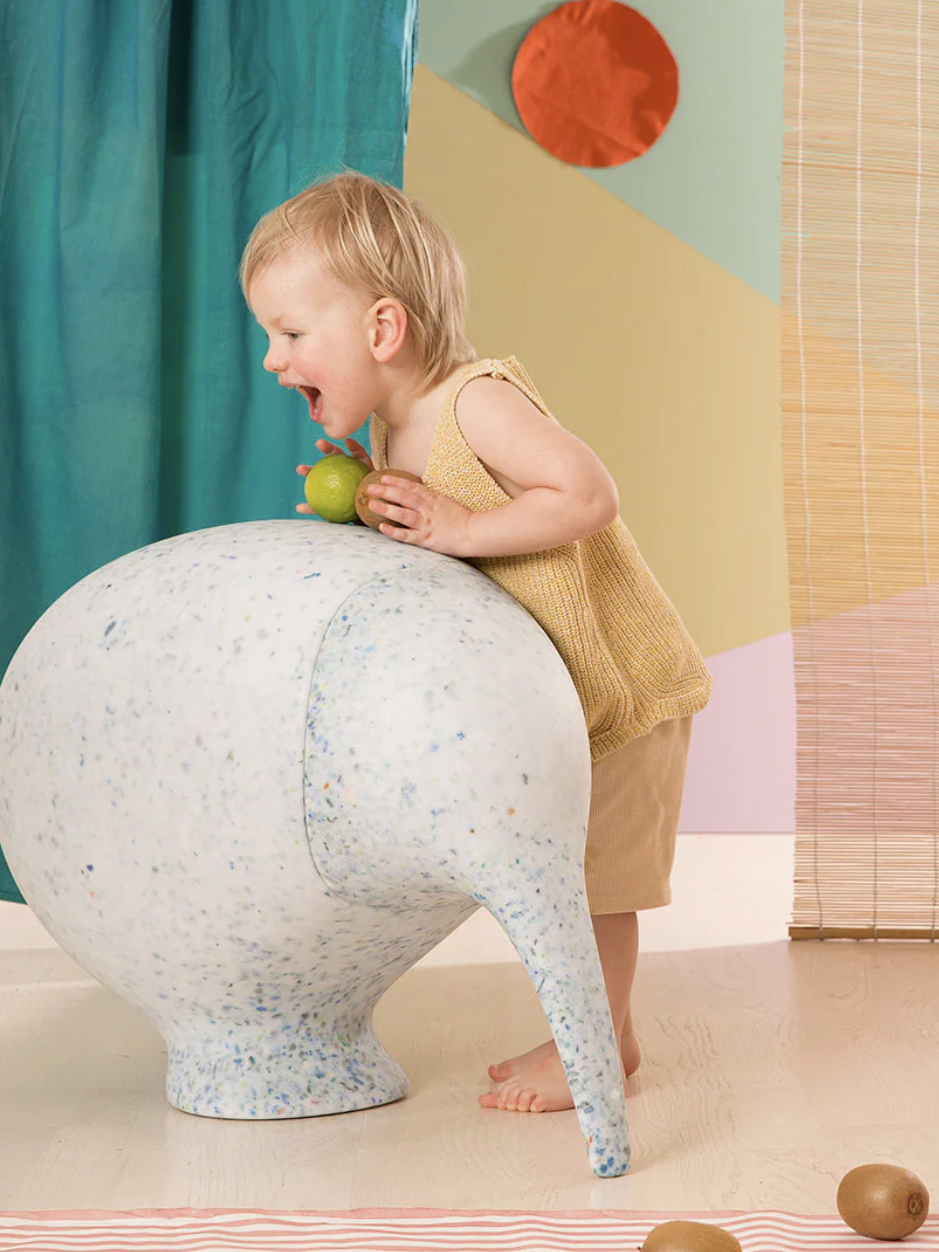 A child leans forward, licking a large, round abstract sculpture while holding a green object. The background features colorful drapes and geometric shapes.