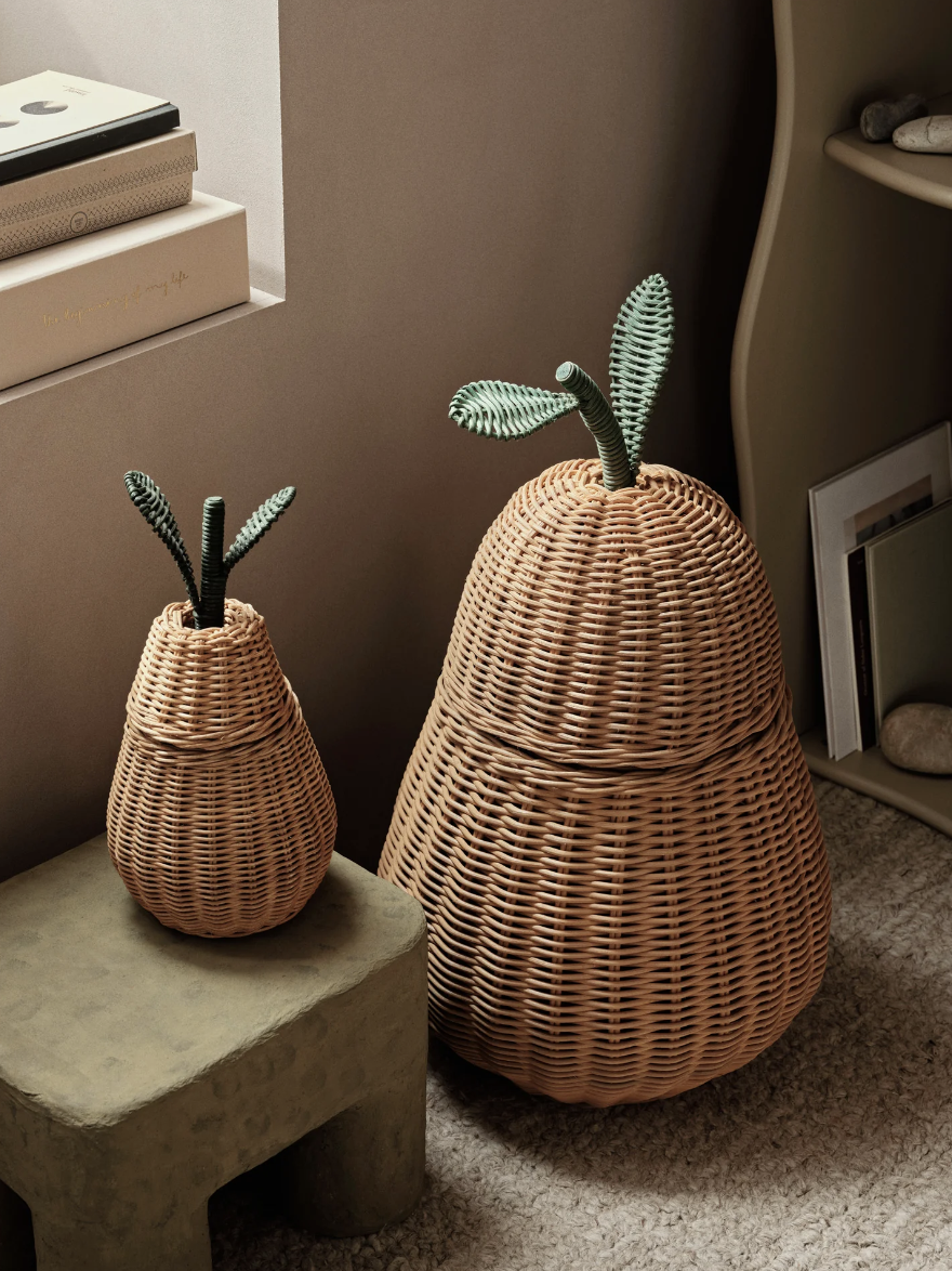 Two woven wicker baskets shaped like pears with green leaves on top, placed on a carpeted floor next to a wall shelf.