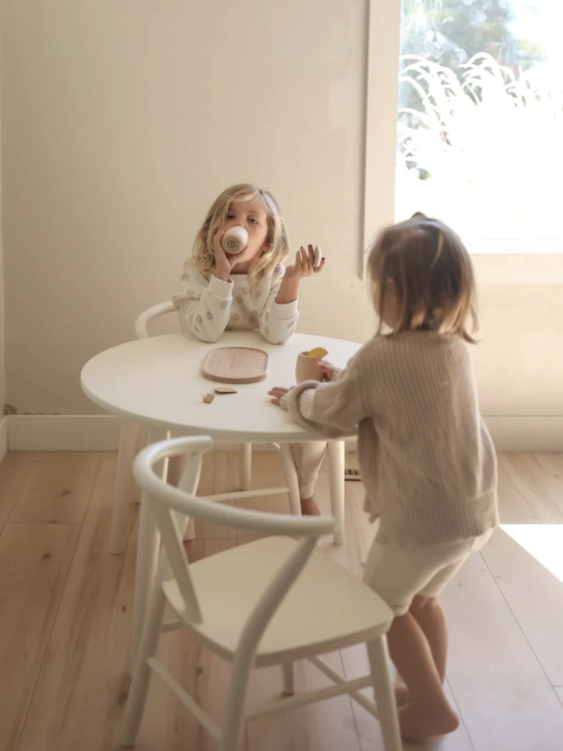 Two children sit at a small white table in a bright room. One child drinks from a cup while the other is standing and playing with a toy.