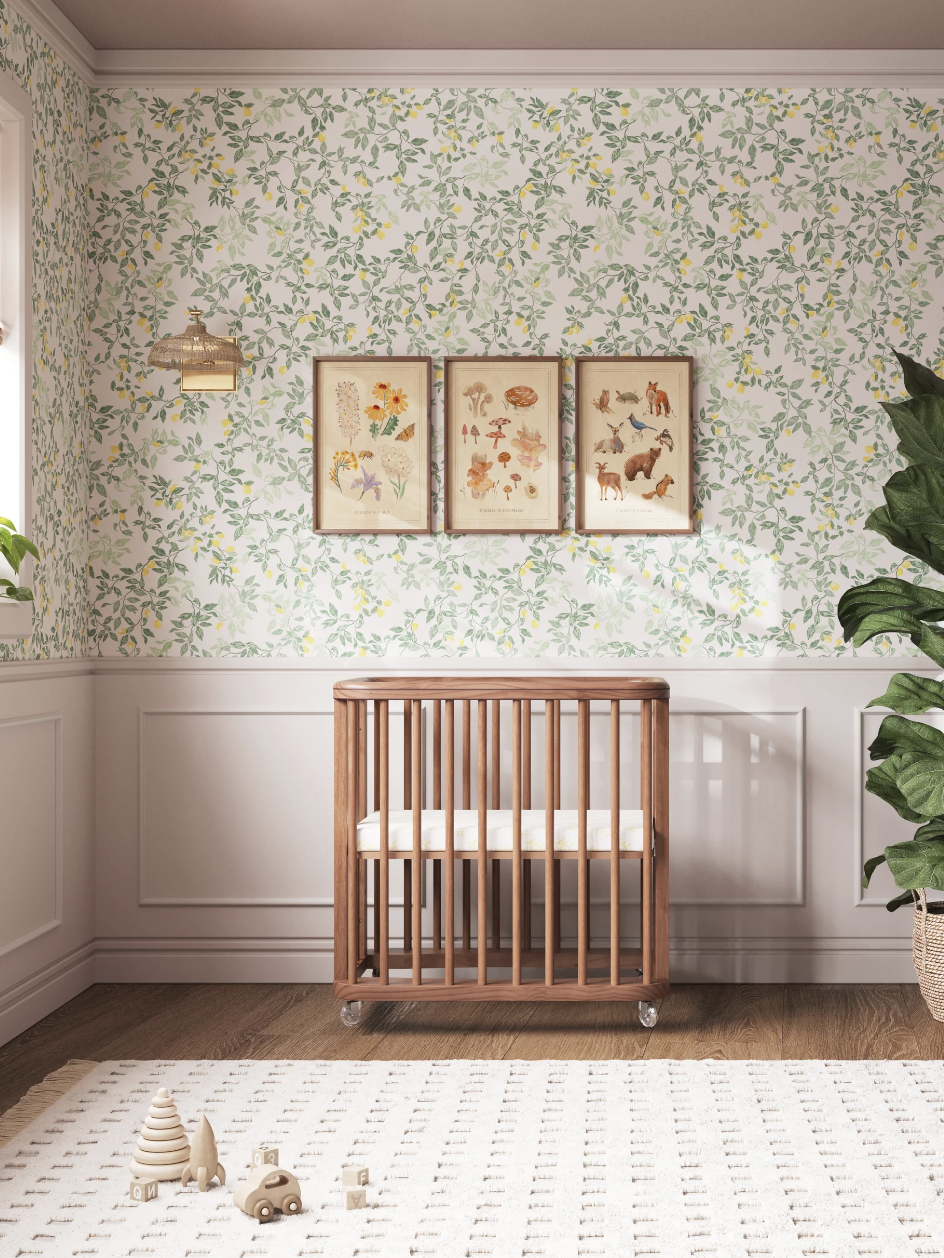 A wooden crib is centered in a nursery with floral wallpaper, three framed art pieces, a potted plant, and a patterned rug.