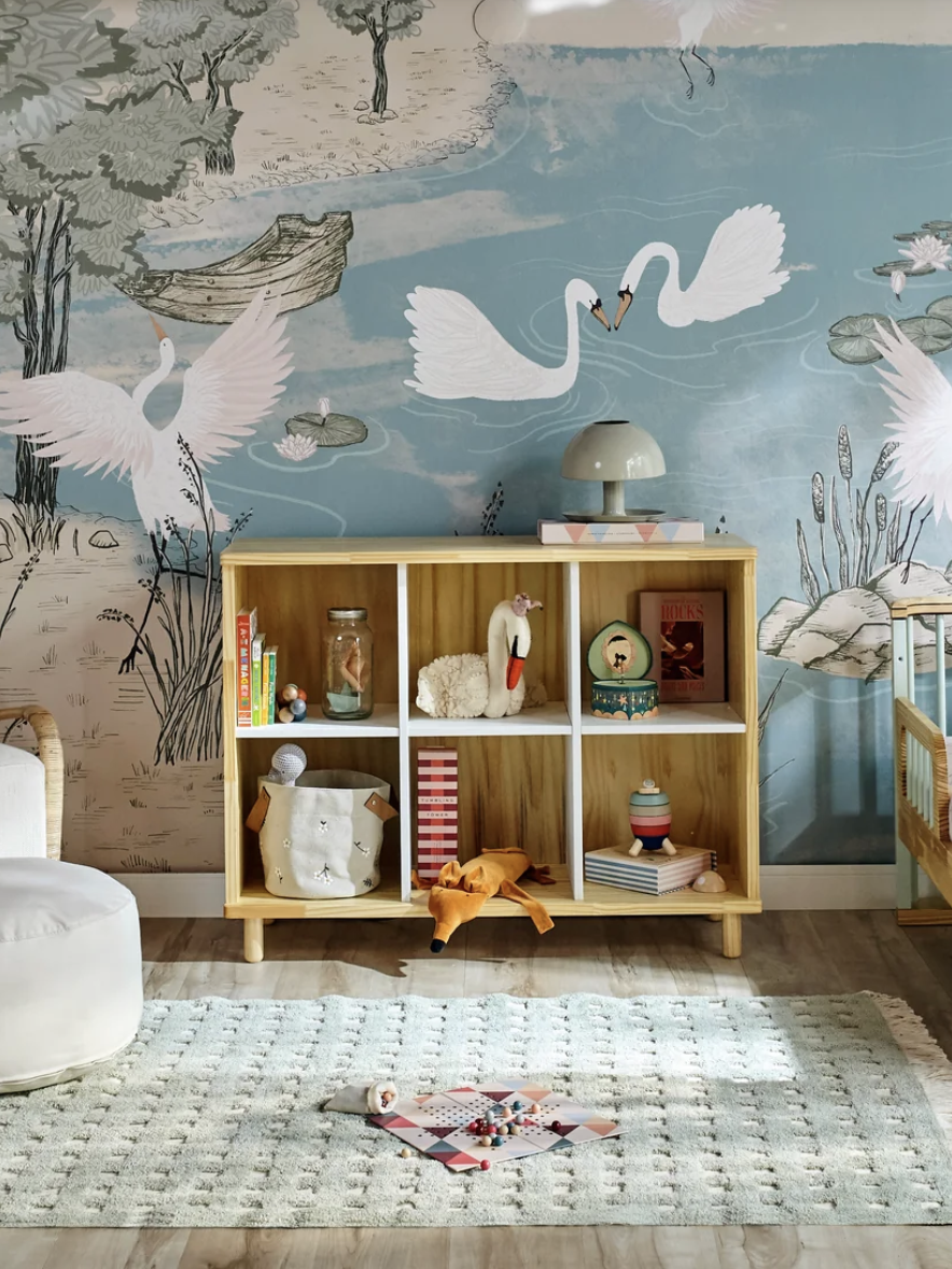 A nursery with swan-themed wallpaper, a crib, bookshelf with toys, and a round ottoman. A soft rug covers the floor. A lamp sits on the shelf.