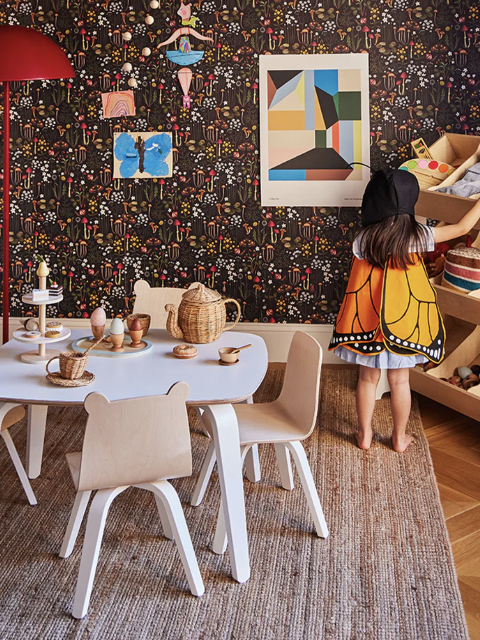 Child in butterfly costume looking at a shelf in a colorful, decorated playroom with a small table and chairs.