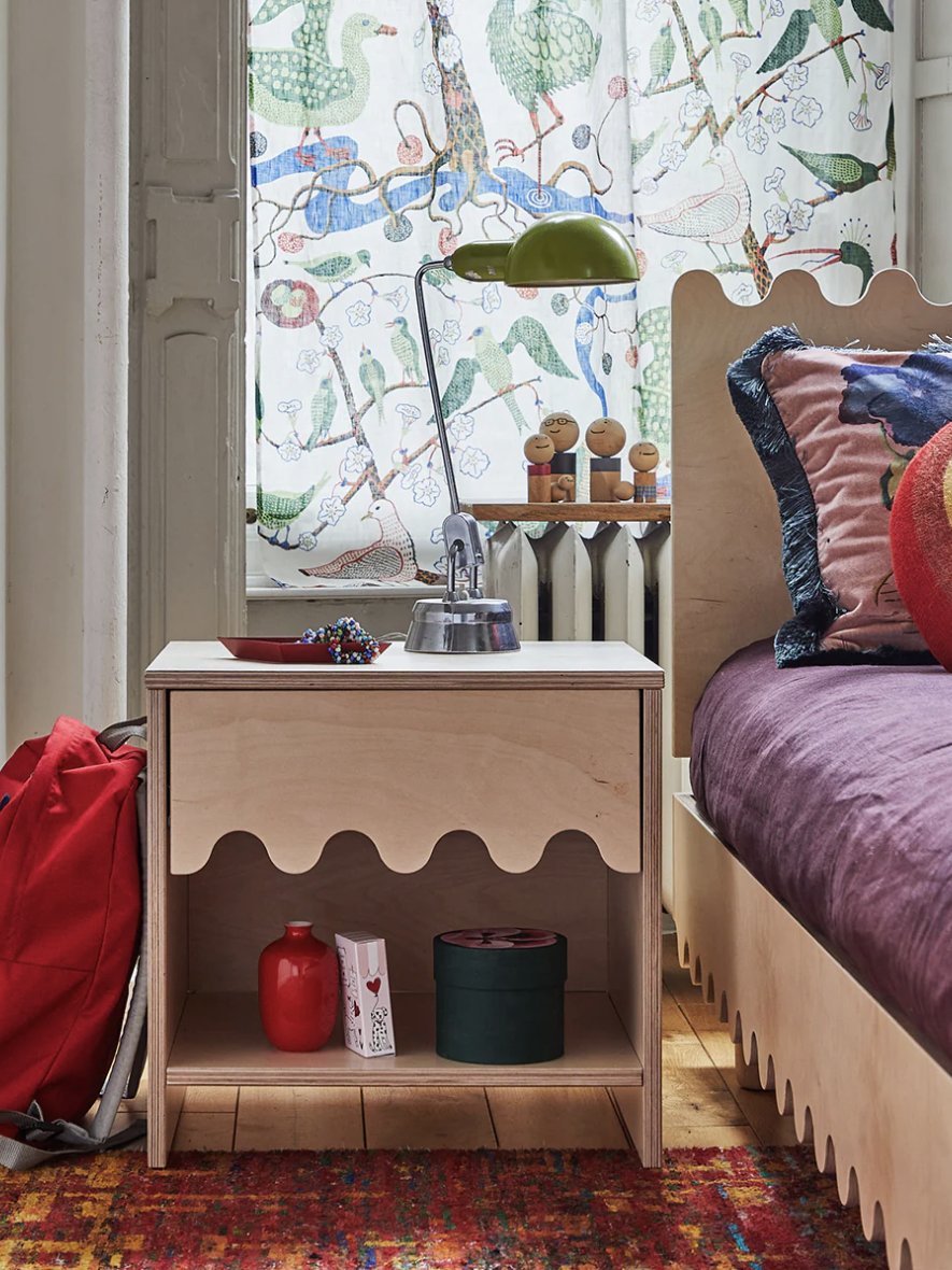 Bedroom scene with a wooden nightstand, a red backpack, and colorful decor. There is a bed with a purple blanket and a multicolored pillow. A window with a bird-patterned curtain is in the background.