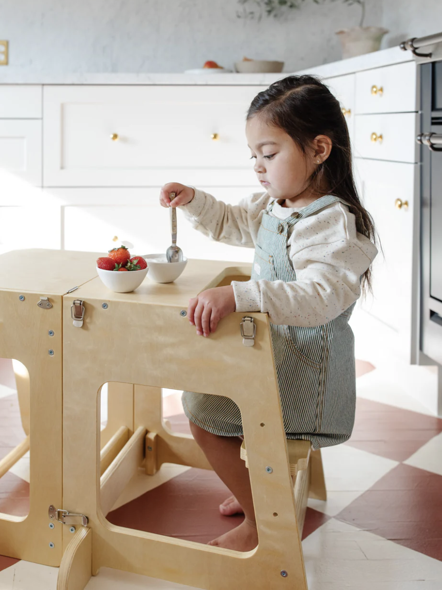 A child sits at a wooden table in a kitchen, eating from a small bowl with a spoon. A bowl of strawberries is on the table.