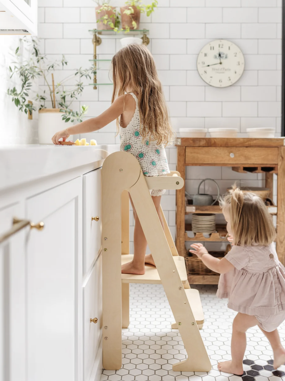 Two young children in a kitchen, one standing on a wooden step stool at the counter, and the other standing nearby on a tiled floor with white cabinetry and a clock on the wall.