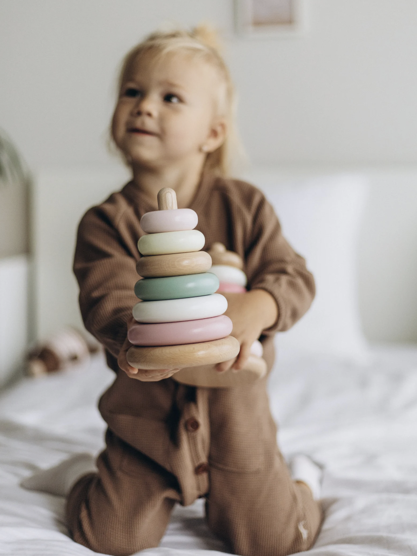 Child kneeling on a bed holding a colorful stack of toy rings, wearing a brown outfit.