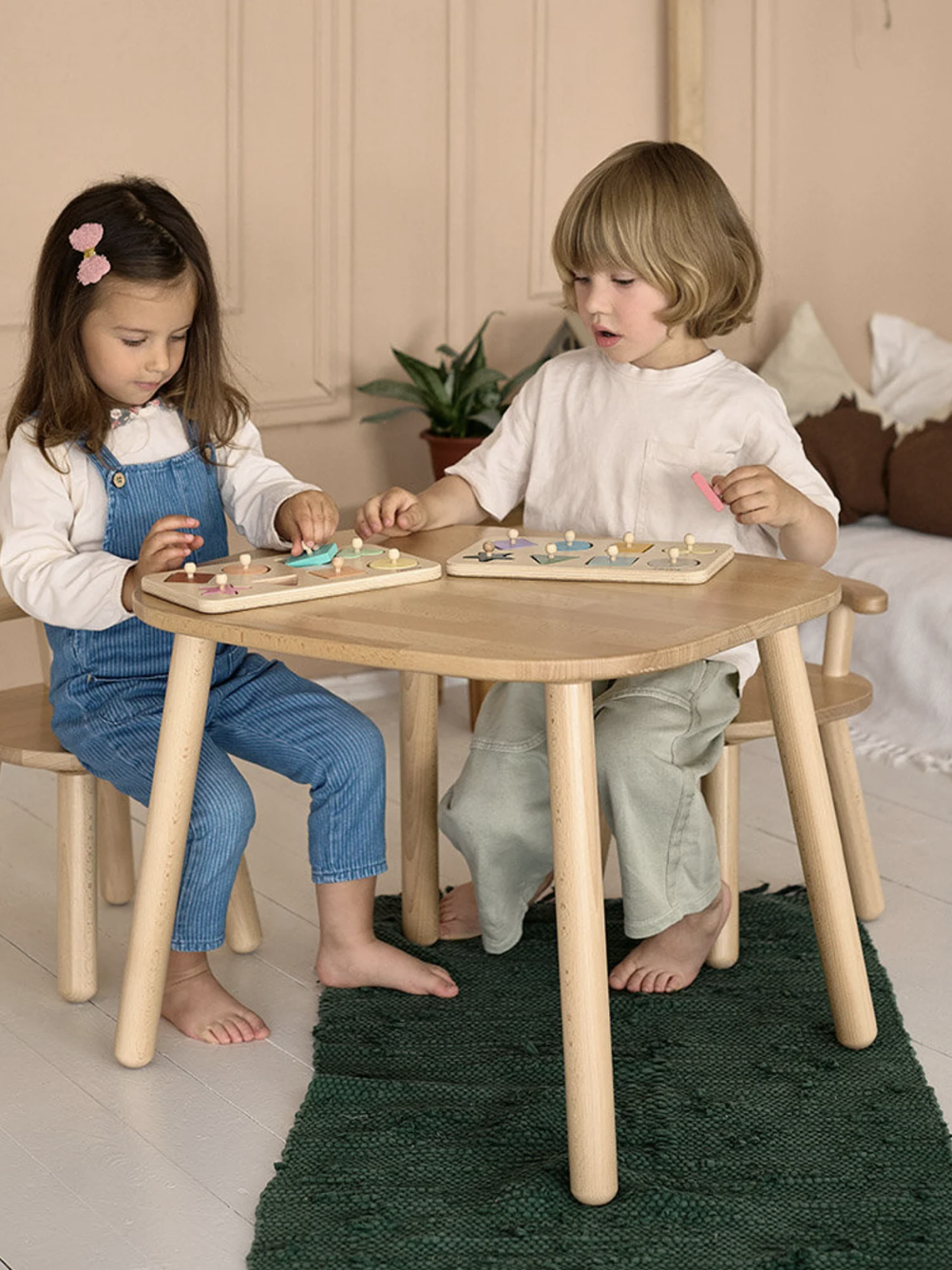 Two children sitting at a wooden table, playing with colorful peg boards. They appear focused and engaged in their activity. There is a plant and pillows in the background.