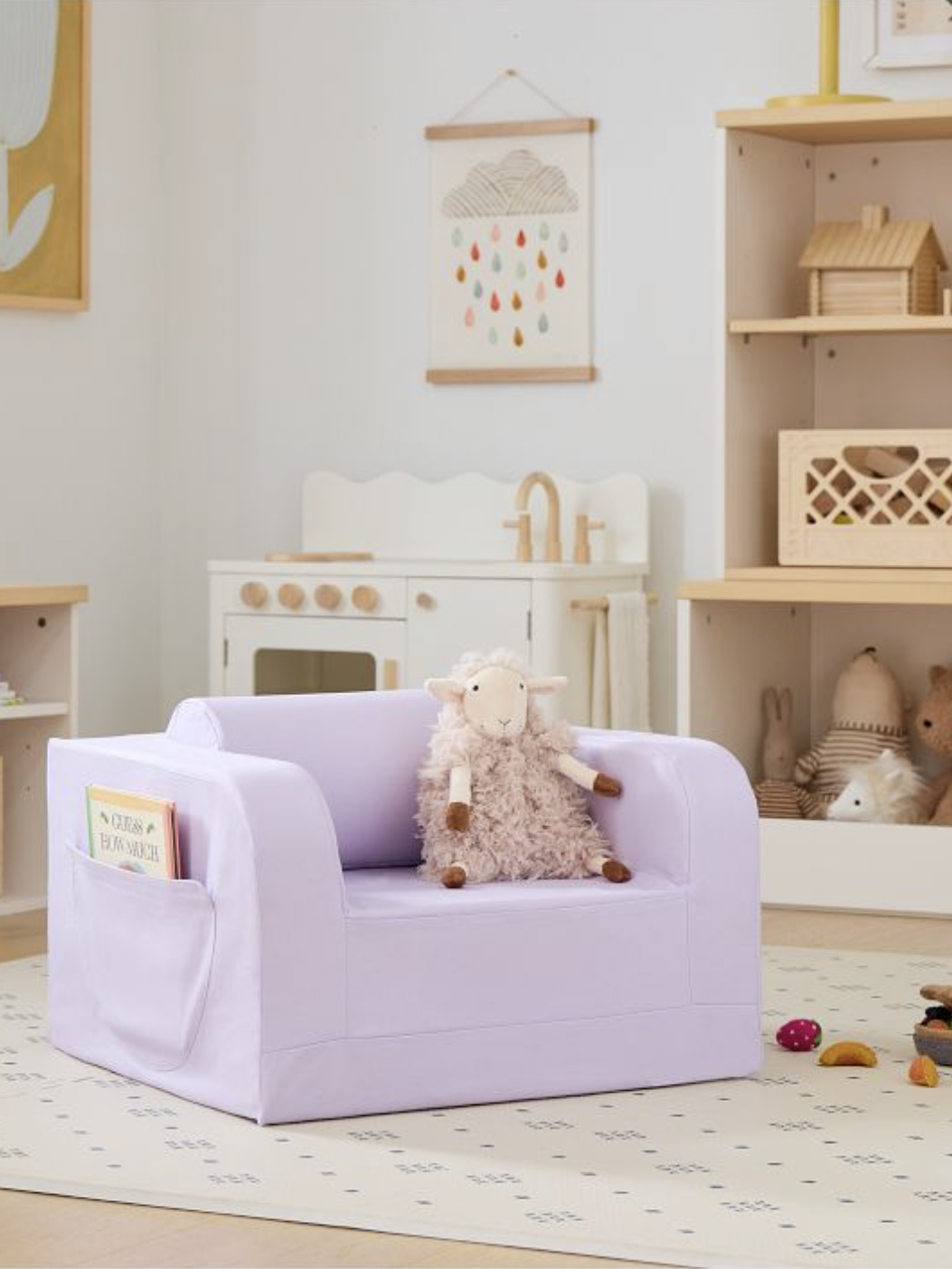 A lavender children's armchair with a plush sheep toy on it is placed in a playroom. A soft book is in the chair's side pocket surrounded by toys and wooden shelves in the background.