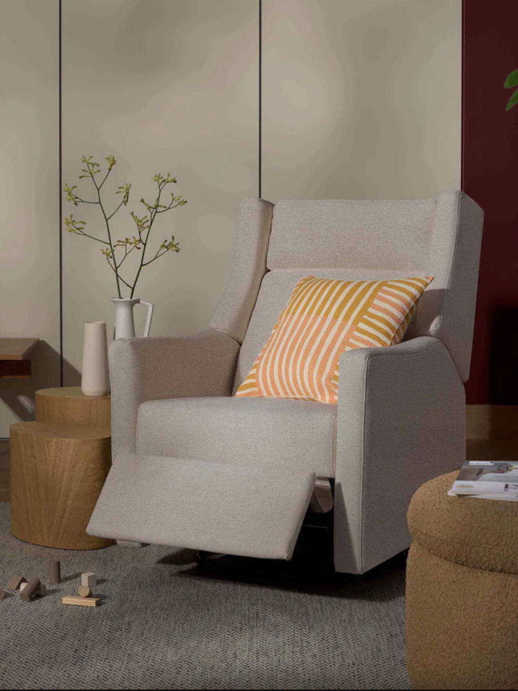 A beige recliner with an orange striped pillow is surrounded by a side table, potted plant, and abstract wall art.