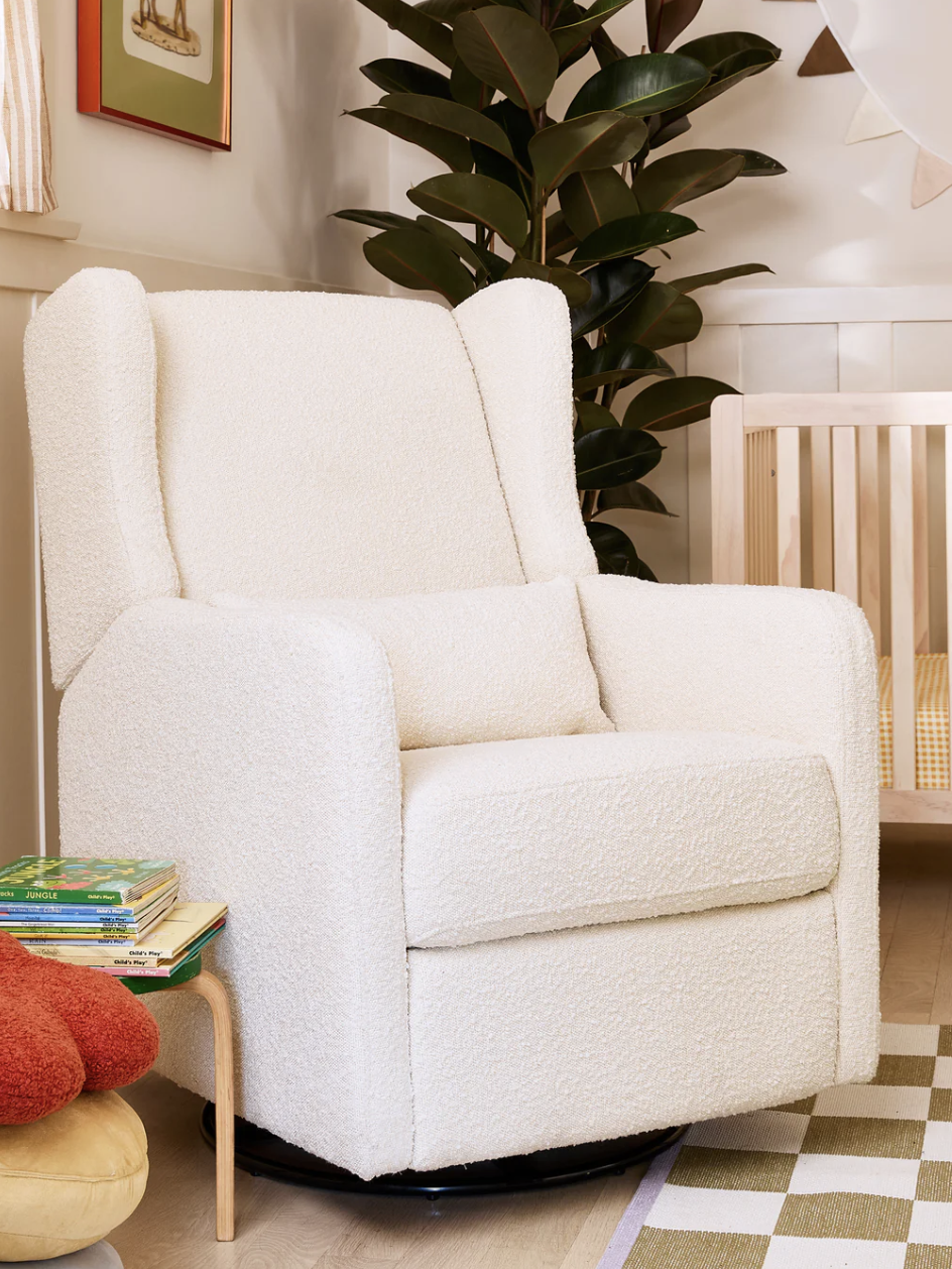 A white upholstered armchair in a cozy, light-filled room, next to a crib and a small table with books and a plush cushion.