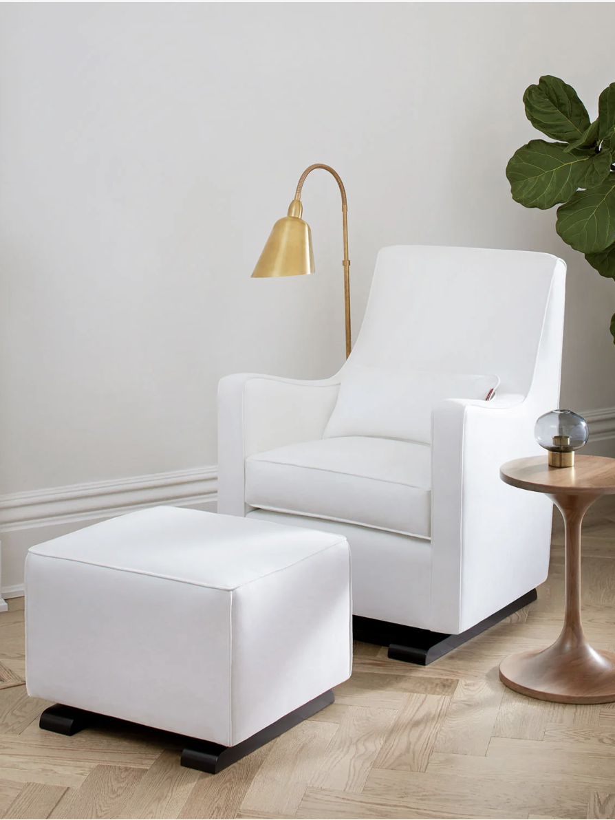 A white armchair with a matching ottoman next to a round wooden table, a brass floor lamp, and a potted plant in a minimalist room.