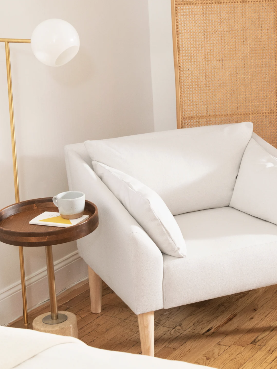 A white cushioned armchair next to a wooden side table with a cup and book, near a wicker room divider on a wooden floor.