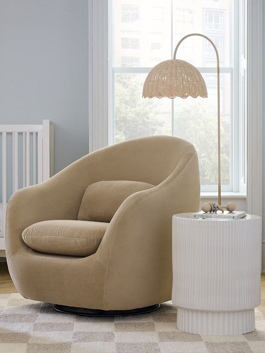 A cozy nursery room with a beige armchair, ottoman, a white crib, a side table, and a standing lamp. A giraffe print hangs on the wall.